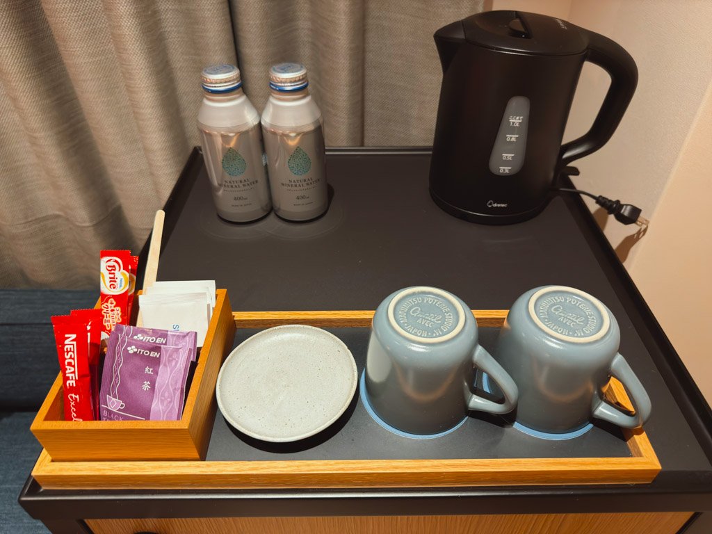 客室内のドリンクセット。コーヒー、紅茶のティーバッグが木製トレイに並び、カップとソーサー、ペットボトル水、電気ケトルが一緒に配置されています。