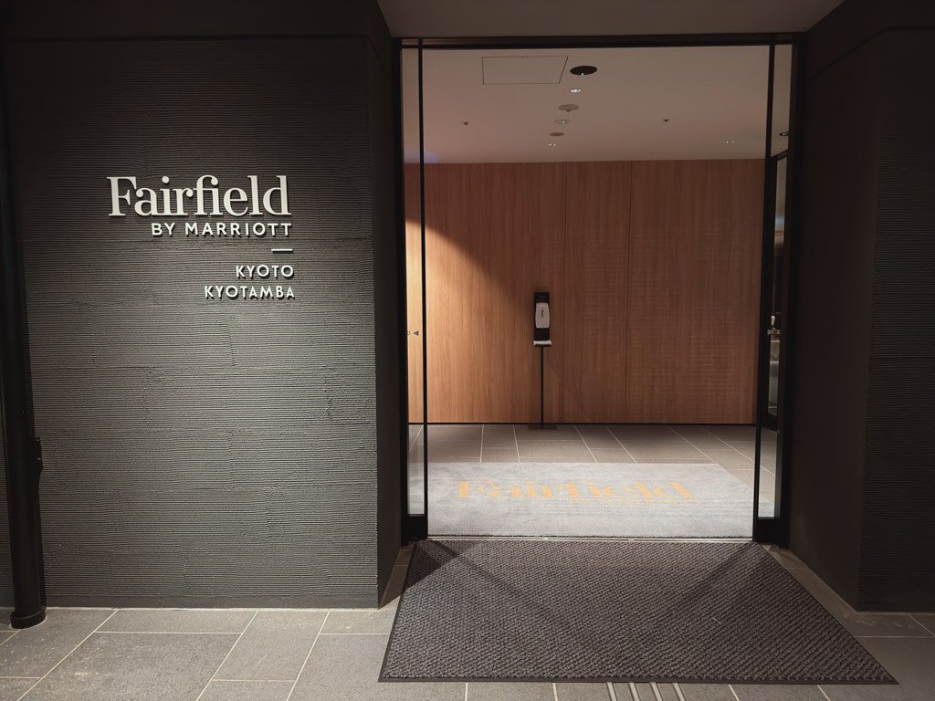 フェアフィールド・バイ・マリオット 京都京丹波のエントランスロゴサイン。落ち着いたグレーの壁にホテル名が明確に表示されており、入口の向こう側には温かみのある木目調の内装と自動手指消毒器が設置されています。