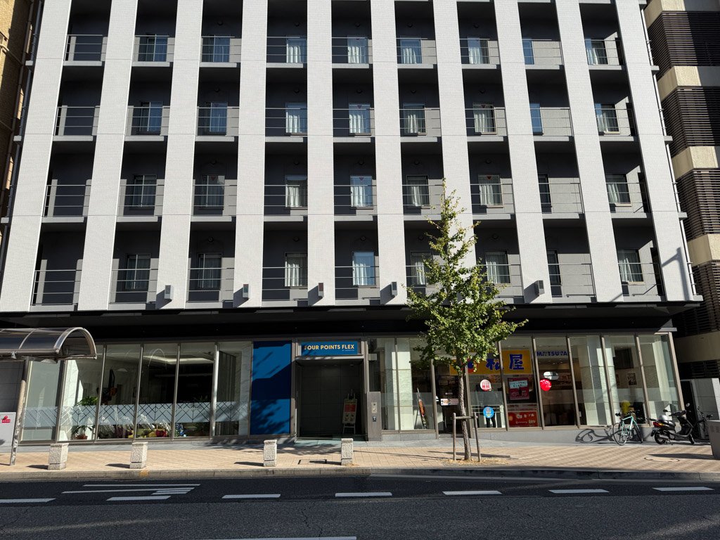 フォーポイント フレックス by シェラトン 神戸三宮の外観。中央にモダンな白いビルがそびえ、左右には異なるデザインの建物が並ぶ。ビルの下には入り口が見え、周囲の街並みが映し出されている。