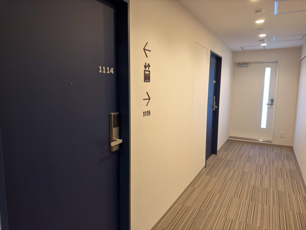 フォーポイント フレックス by シェラトン 神戸三宮の客室フロア廊下。青いドアの部屋番号「1114」が目立ち、清潔感のある廊下と案内サインが見える。