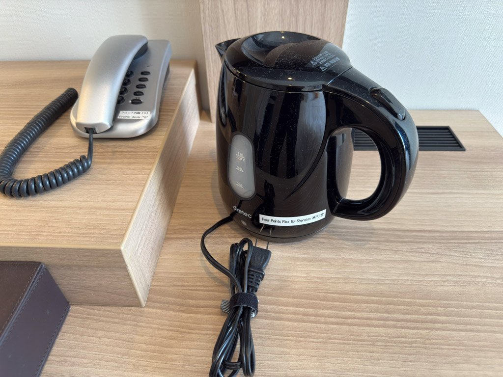フォーポイント フレックス by シェラトン 神戸三宮の客室内に設置された黒い電気ケトルと電話機。木製のデスクの上にコンパクトにまとめられている。