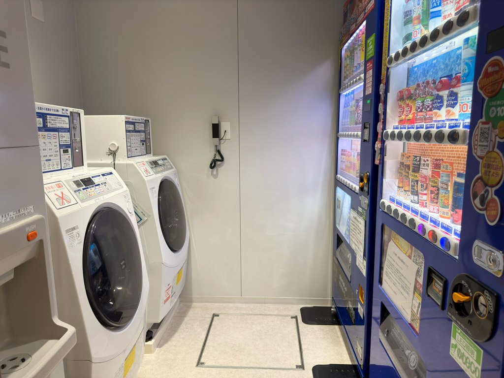 フォーポイント フレックス by シェラトン 神戸三宮の1階に設置された自動販売機と洗濯乾燥機。明るいスペースに並ぶ機械が、便利な設備を提供している。