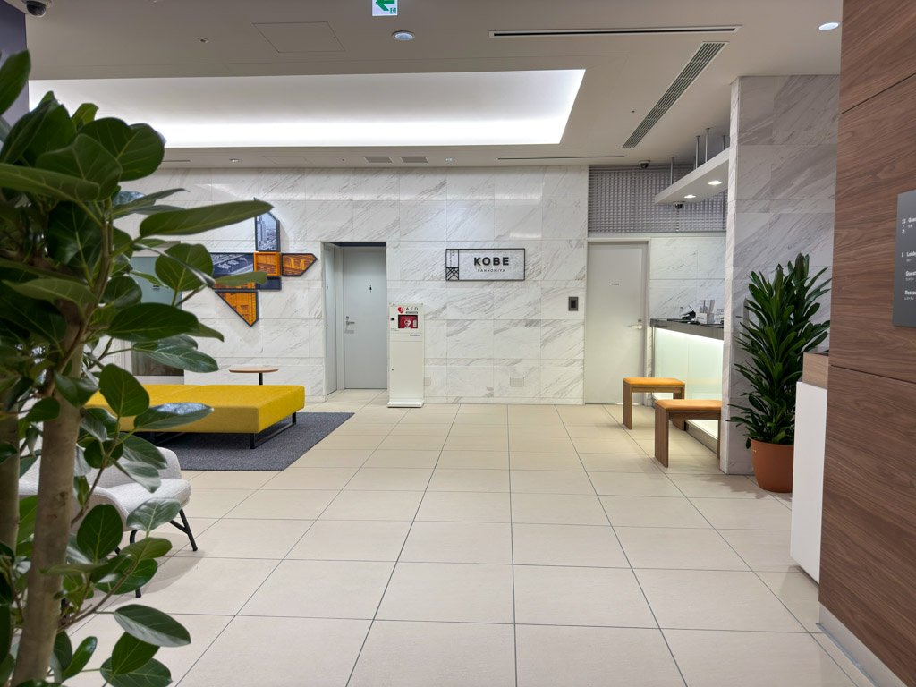 フォーポイント フレックス by シェラトン 神戸三宮のロビー全景。植物が配置されたモダンな空間には黄色いソファやテーブルがあり、奥には「KOBE SANNNOMIYA」のサインやレセプションカウンターが見える。