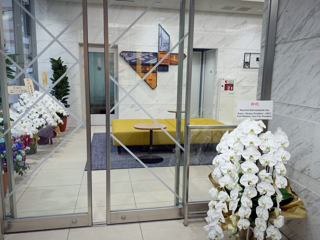 フォーポイント フレックス by シェラトン 神戸三宮のエントランス内。オープン初日を祝う白い胡蝶蘭の花が飾られ、奥には黄色いソファとテーブルが置かれたモダンなロビーが見える。