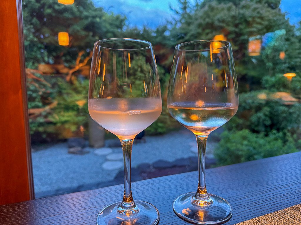  奈良の日本酒「みむろ杉」を注いだワイングラス2つ、和風庭園を背景にした美しいシーン  Two wine glasses filled with Nara’s sake “Mimuro Sugi,” set against a backdrop of a traditional Japanese garden 