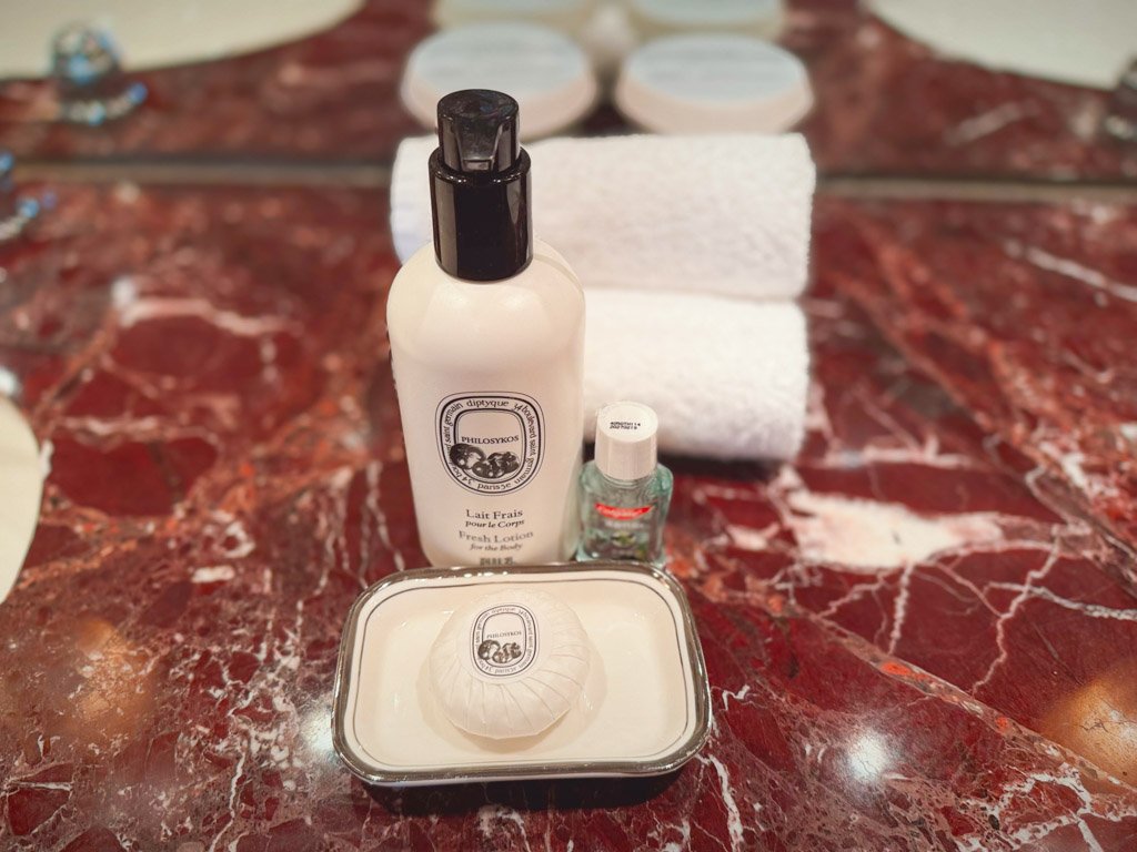  バスルームの大理石カウンターの上に並べられたディプティック（Diptyque）のアメニティセット。ボディローション、石鹸、ミニサイズのボトルがセットされており、上品で贅沢な雰囲気が漂っています。清潔感のあるタオルも並んでおり、ラグジュアリーな滞在を演出しています。 