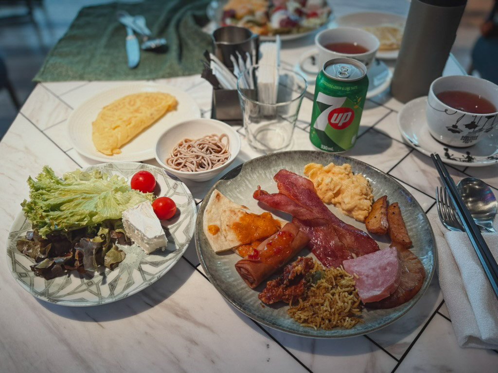  Wマカオ・スタジオシティの朝食ビュッフェの一例で、さまざまな料理が並べられています。オムレツ、そば、サラダ、ベーコン、スクランブルエッグ、焼き野菜などが盛られており、朝食に豊富な選択肢があることがわかります。また、7UPと紅茶も飲み物として用意されており、カジュアルでリラックスできる朝食のひとときが楽しめそうです。 