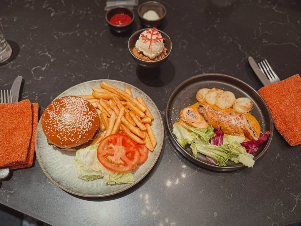  インルームダイニングで提供された料理。左側にはセサミバンズのハンバーガーとポテトフライ、レタスとトマトの付け合わせがあり、右側にはチキンのグリルとサラダが添えられています。中央にはデザートもあります。オレンジ色のナプキンがアクセントになっており、テーブル全体がスタイリッシュで落ち着いた雰囲気です。 
