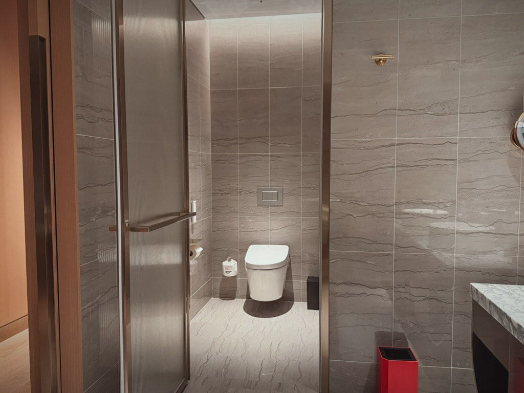  トイレの写真です。ガラスの仕切りドアがあり、落ち着いたデザインのタイルが壁に施されています。シンプルなトイレ設備に加え、赤いゴミ箱がワンポイントとしてアクセントになっています。 