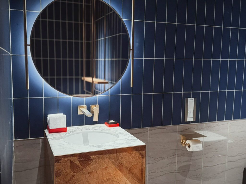  来客用トイレの洗面台エリア。円形のミラーが壁に映え、青いタイルとゴールドの蛇口が高級感を演出しています。洗面台は大理石調で、タオルと小物が整然と配置されています。 