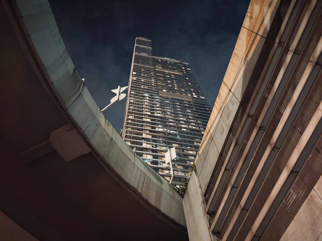  高層ビルを下から見上げた夜景の写真。近代的な建築物が特徴的で、手前には道路や構造物がフレームに含まれている。 