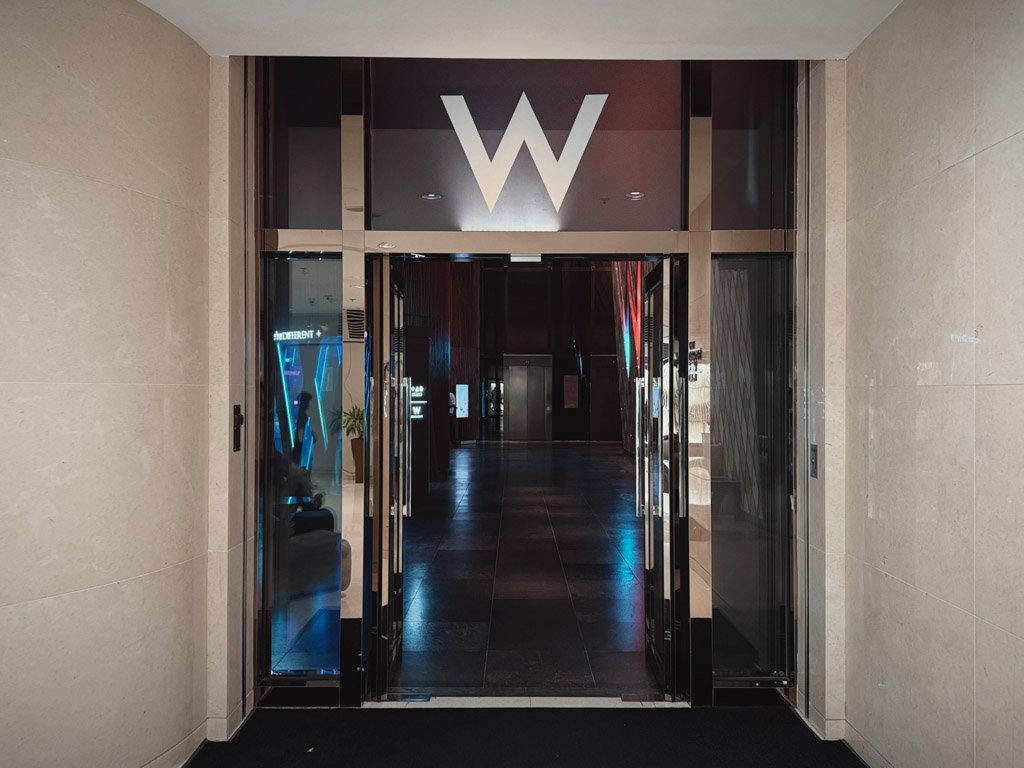  W香港の九龍駅側の入り口。ガラス扉の上部には大きな「W」のロゴが光り、モダンで洗練されたデザインのエントランスが映っている。 