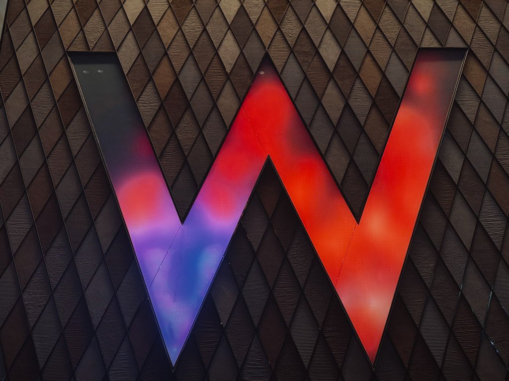 W香港の象徴的な「W」のロゴ。紫と赤のライトで照らされ、クラブのような華やかな雰囲気を演出している。背景にはダイヤ柄の壁が特徴的に配置されている。 