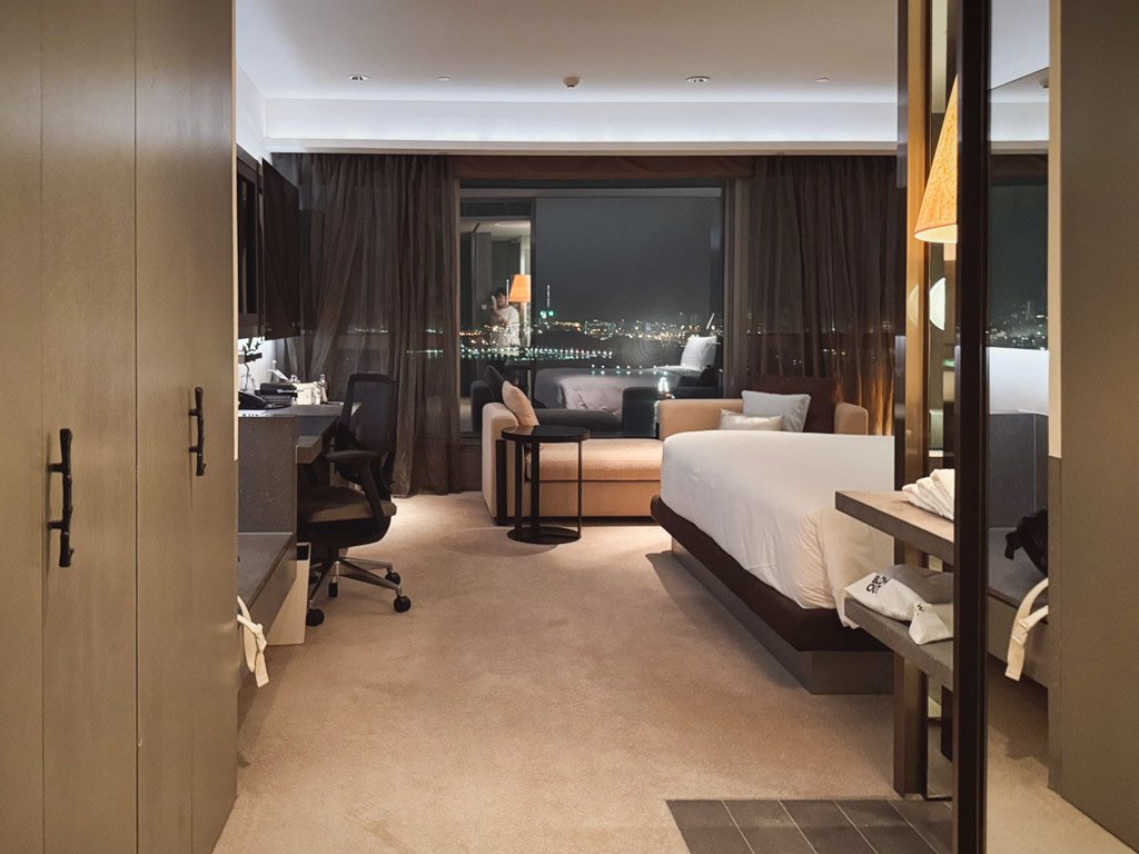  W香港の客室内部。広々としたモダンなデザインの部屋には大きなベッド、デスク、ソファが配置され、窓の外には美しい夜景が広がっている。 