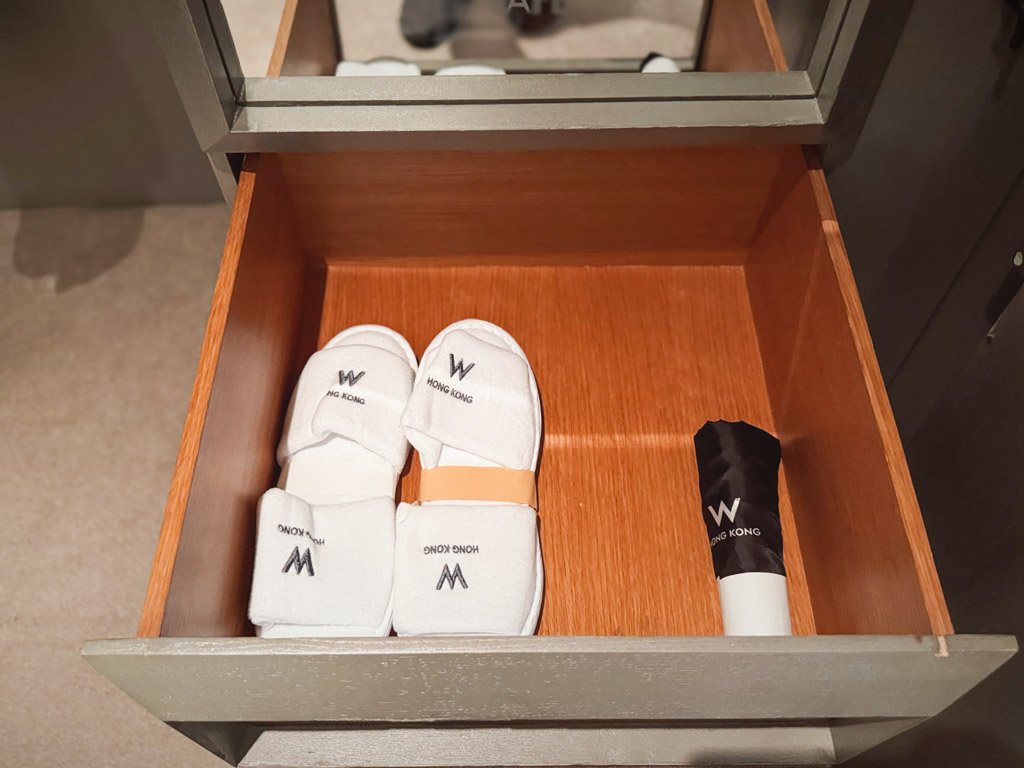  W香港の客室内、引き出しに収納されたスリッパと買い物袋。白いスリッパには「W HONG KONG」のロゴがデザインされており、黒い傘もブランドロゴ入りでスタイリッシュにまとめられている。 