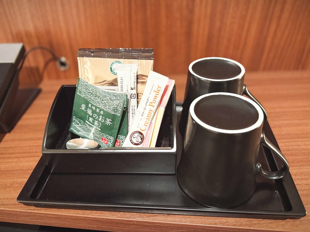 フォーポイント フレックス by シェラトン 横浜駅西の客室に用意されたお茶セット。黒いカップ2つと共に、緑茶、コーヒー、クリーミーパウダーなどがトレーに整然と並べられている。