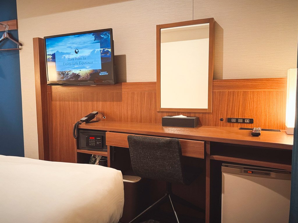 フォーポイント フレックス by シェラトン 横浜駅西の客室内にあるデスクエリア。壁掛けテレビ、鏡付きのデスク、セーフティボックス、冷蔵庫が備え付けられており、機能的で快適な作業スペースが提供されている。