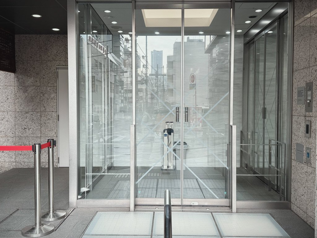 フォーポイント フレックス by シェラトン 横浜駅西のエントランスのガラス扉。透明な自動ドアが建物内を見せ、シンプルで洗練されたデザインが特徴。周辺の街並みもガラス越しに見える。