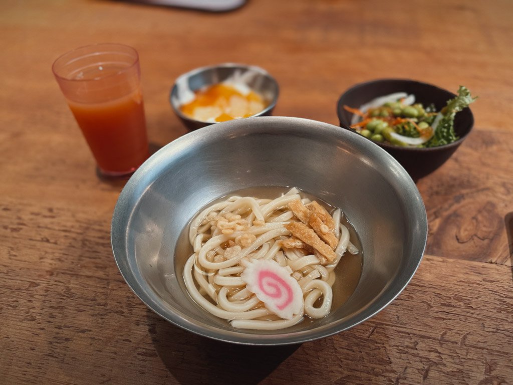 モクシー大阪本町で提供された朝食のうどんセット。ステンレスの器に盛られたうどんには、かまぼこと揚げがトッピングされている。背景には、グレープジュース、小鉢に入ったフルーツ、サラダが並べられている。