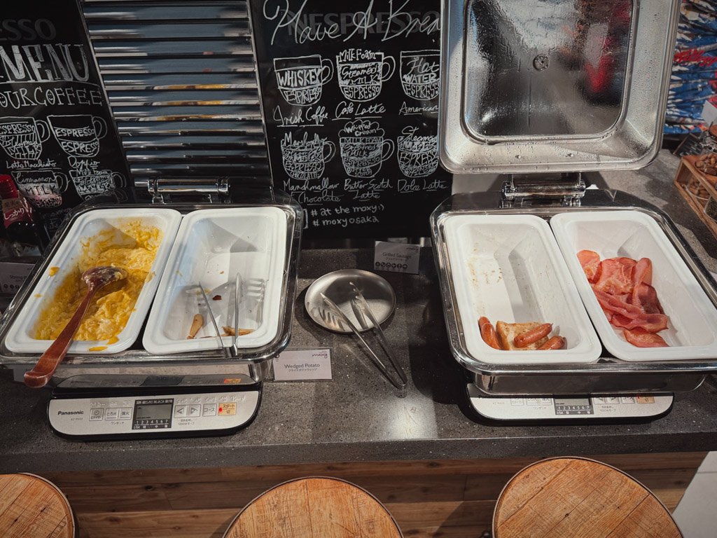 モクシー大阪本町の朝食ビュッフェエリア。スクランブルエッグ、ベーコン、ソーセージが温かいトレイに並べられている。背面には黒板に書かれたドリンクメニューが見える。