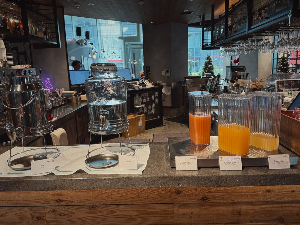 モクシー大阪本町の朝食コーナーにあるドリンクステーション。冷たいウォーターサーバー、グレープフルーツジュース、オレンジジュース、アップルジュースが整然と並べられ、ゲストが自由に利用できる。