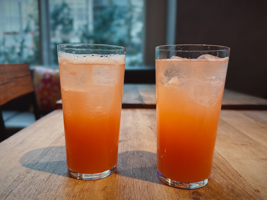 モクシー大阪本町のレストランで提供された2杯のモクシーカクテル。透明なグラスに鮮やかなオレンジ色のカクテルが注がれ、氷が入っている。背景には木製のテーブルと窓際の景色が見える。