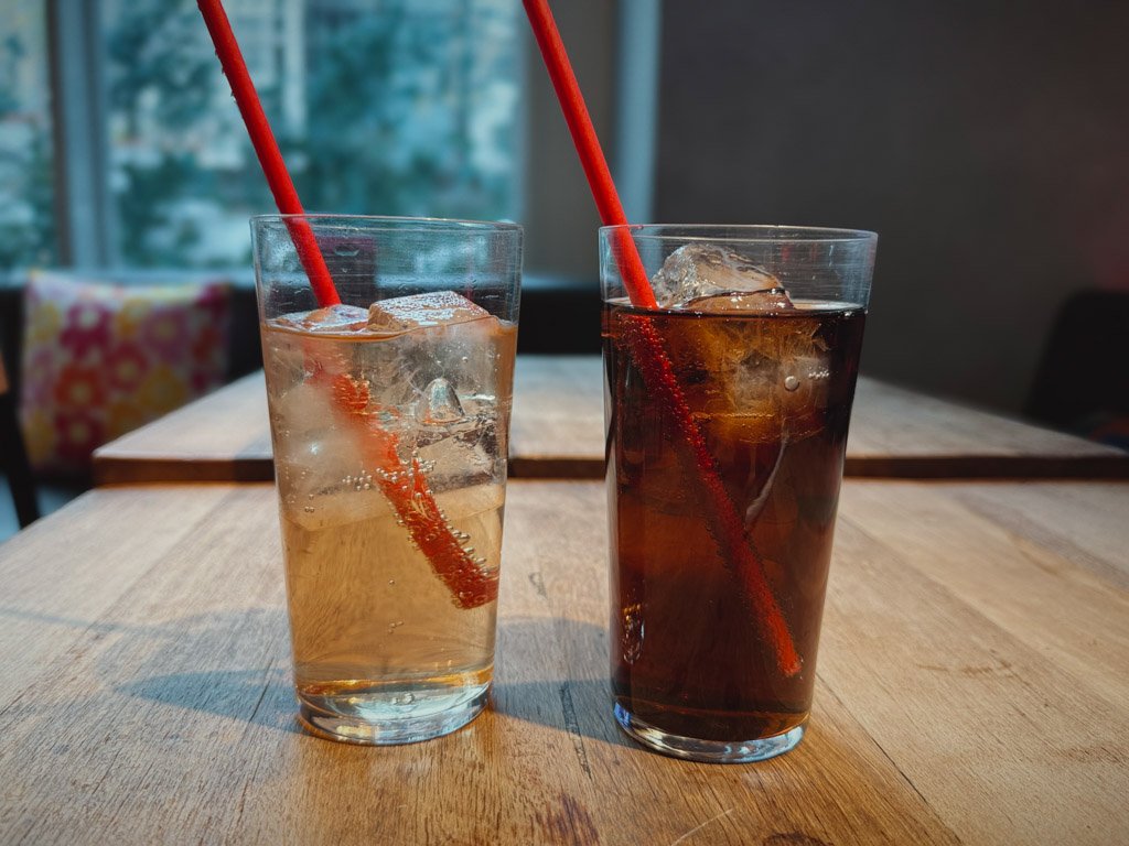 モクシー大阪本町のレストランで提供されるアイスドリンク2杯。透明なグラスに入った炭酸飲料と、赤いストローがささっている。背景には木製のテーブルと窓際の席が見える。