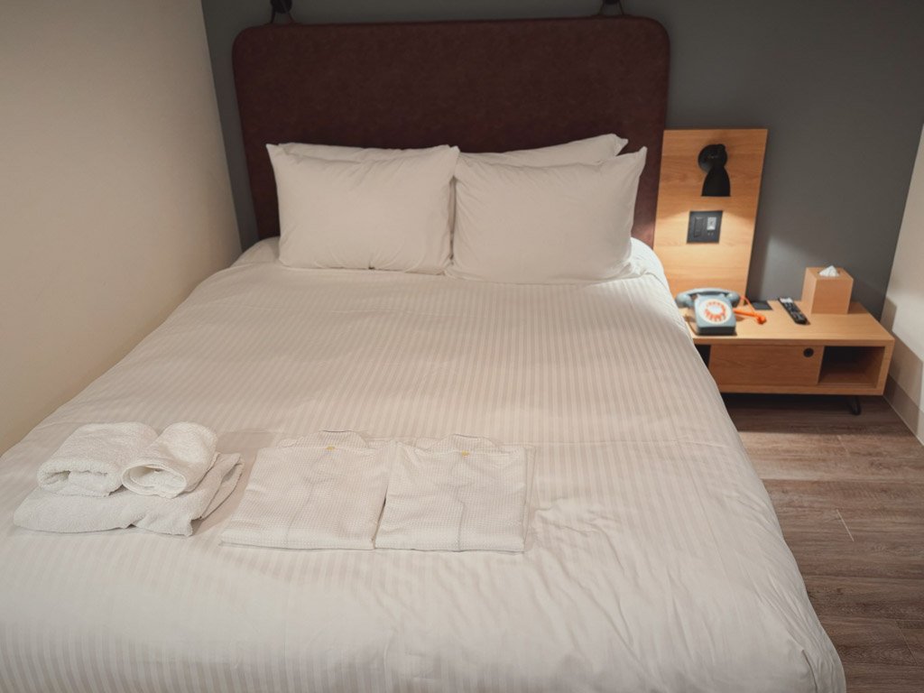 モクシー大阪本町の客室内にある整えられたダブルベッド。ベッドの上にはタオルと部屋着が丁寧に用意されています。横には木製のサイドテーブルがあり、レトロな電話とティッシュボックスが置かれています。