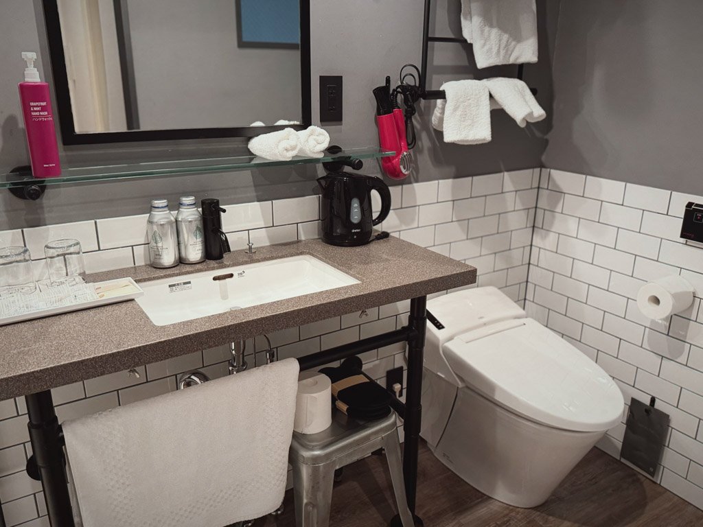 モクシー大阪本町のバスルーム内。洗面台とその横に配置されたトイレが写っています。モダンなタイルデザインが施され、電気ポットやタオルが整然と並べられています。シンプルながら機能的な設備が特徴です。