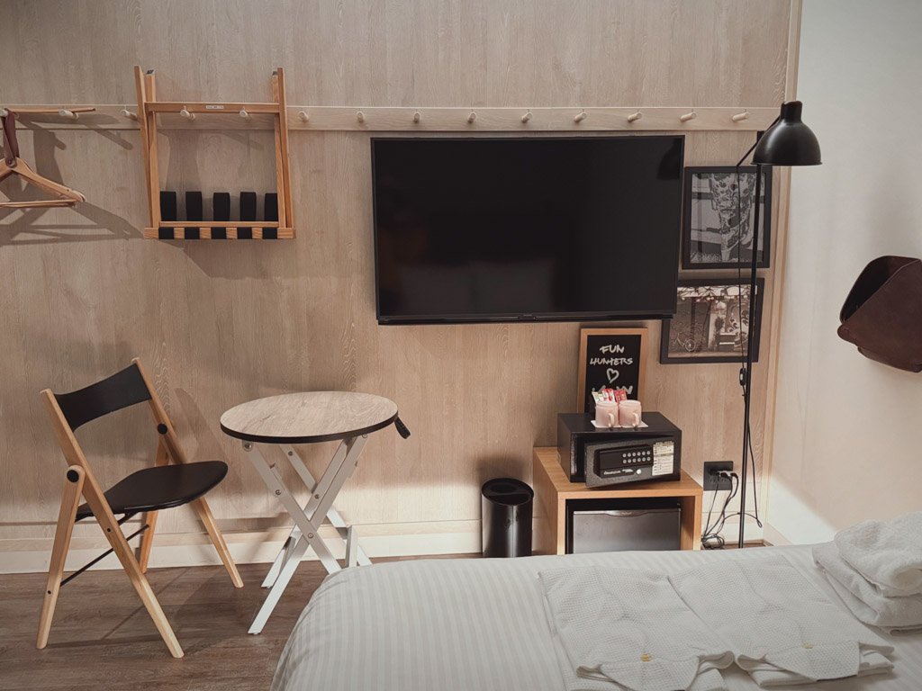 モクシー大阪本町の客室内の一部。壁に取り付けられたフラットスクリーンテレビとハンガーラック、簡易的な木製のテーブルと椅子が見えます。テレビの下には金庫と収納棚が配置され、横にはモダンなフロアランプが設置されています。