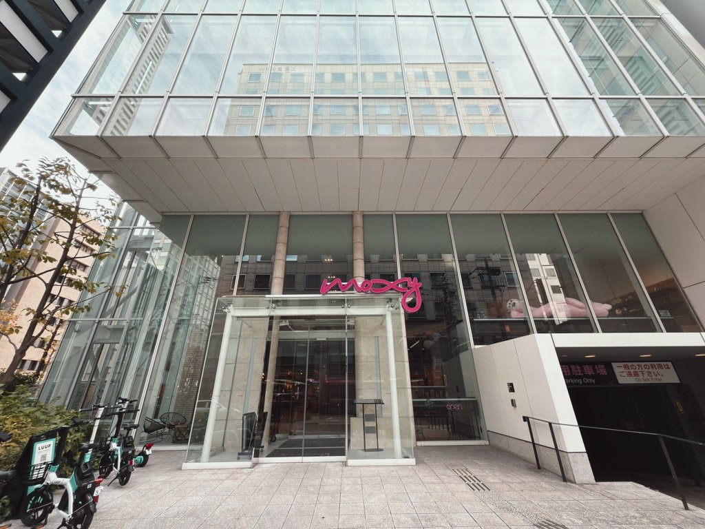 モクシー大阪本町のエントランス。全面ガラス張りの近代的な建物に、特徴的なピンクの「moxy」ロゴが映えています。エントランス付近には電動スクーターが並べられ、都会的でスタイリッシュな雰囲気を演出しています。