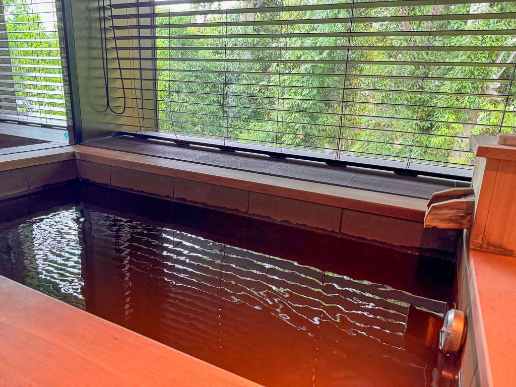  温泉付きの風呂場。窓の外に緑が広がっています。  Onsen bath area with greenery visible outside the window. 