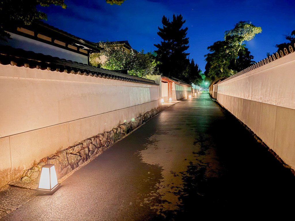  夜のホテル「紫翠」の通路、白壁に沿って静かにライトアップされた落ち着いた雰囲気の通路  Night pathway at the hotel “Shisui,” featuring a serene atmosphere with softly lit white walls along the quiet walkway 