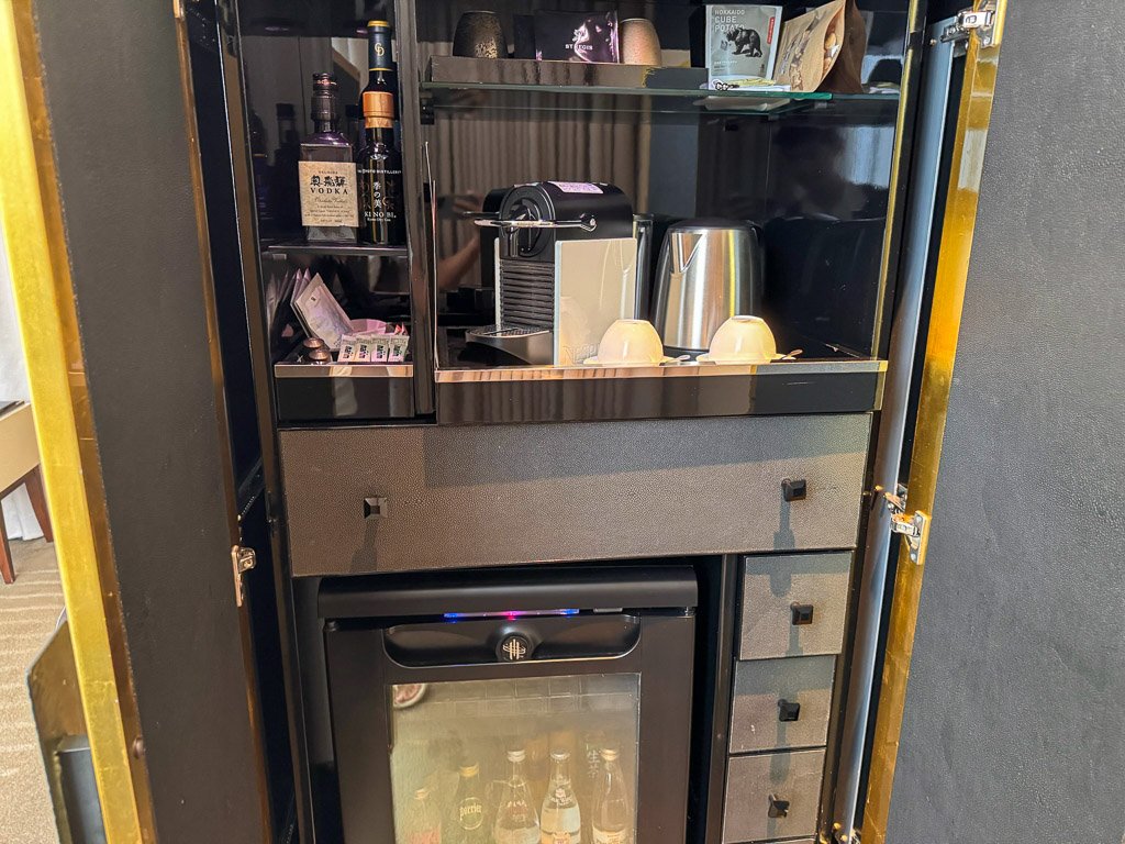 セントレジス大阪の客室内にあるミニバーとコーヒーステーション。洗練されたキャビネット内にきちんと整えられており、ミニバーには高級飲料が揃っています。コーヒーステーションにはネスプレッソマシンとティー用のケトルがあり、滞在中のゲストにラグジュアリーと利便性を提供します。  The in-room minibar and coffee station at The St. Regis Osaka, neatly arranged within a sleek cabinet. The minibar includes a selection of premium beverages, while the coffee station features a Nespresso machine and a kettle for making tea, offering guests both luxury and convenience during their stay.  