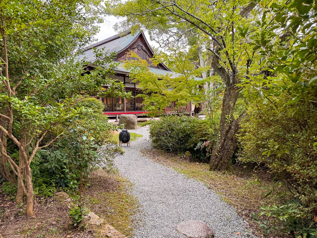  緑に囲まれた石畳の小道が伝統的な日本建築へと続く、静かな庭園の一角  A stone-paved path surrounded by greenery leading to a traditional Japanese building, nestled in a tranquil garden 