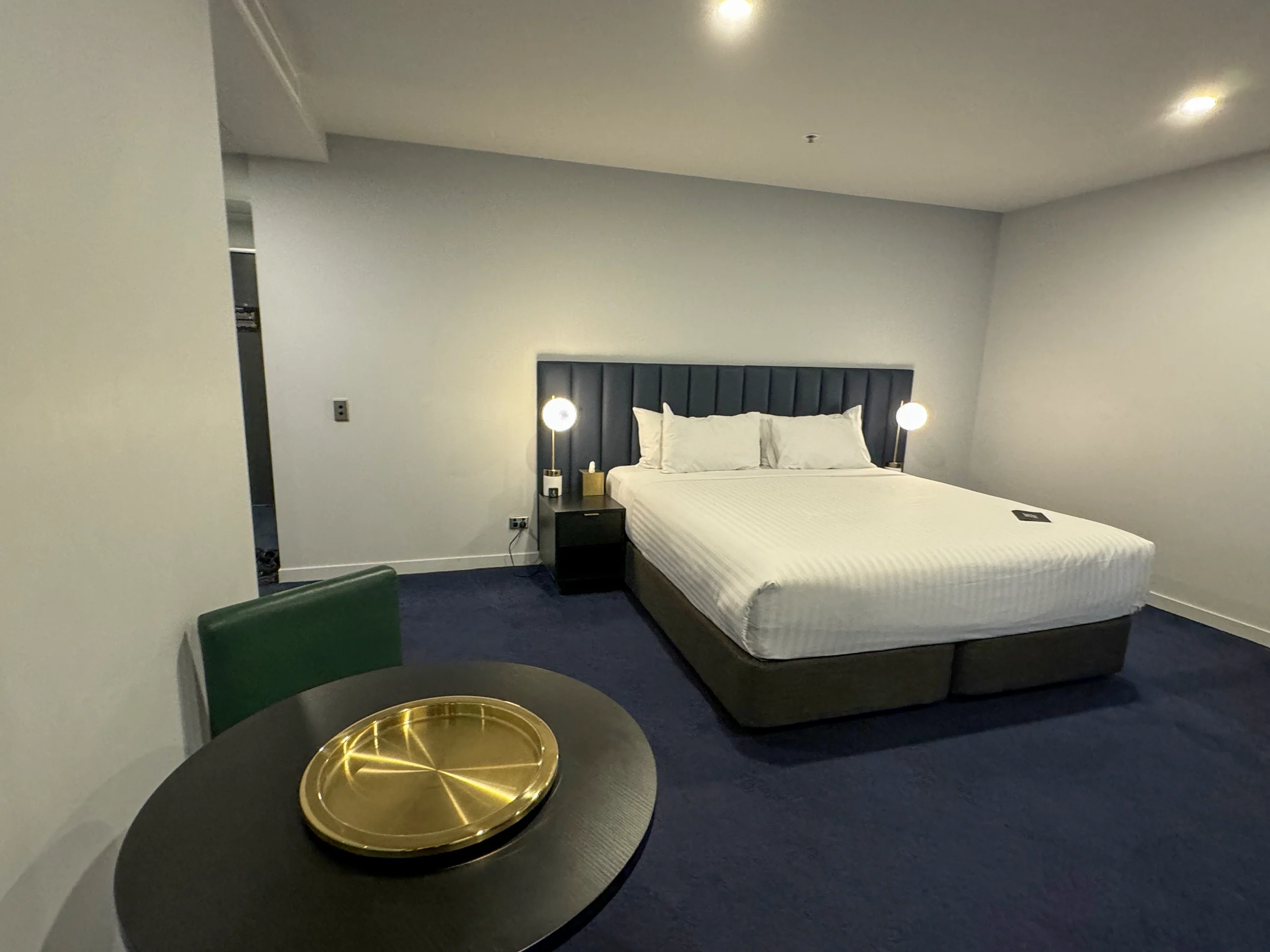 A round table and green chairs, with a white bed beyond.