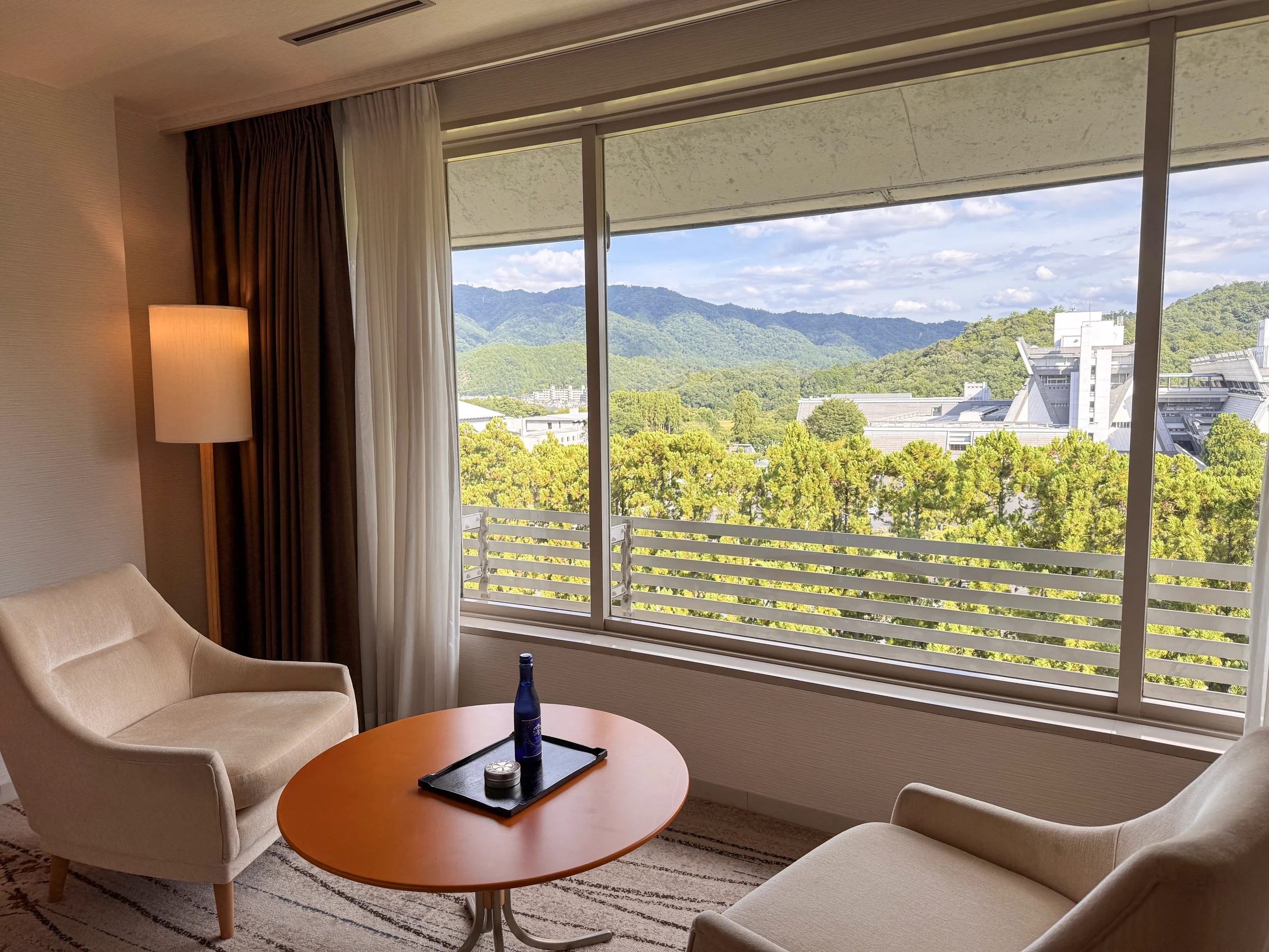 A room at The Prince Kyoto Takaragaike, with chairs and a round table by the window overlooking northern Kyoto’s greenery and hills
